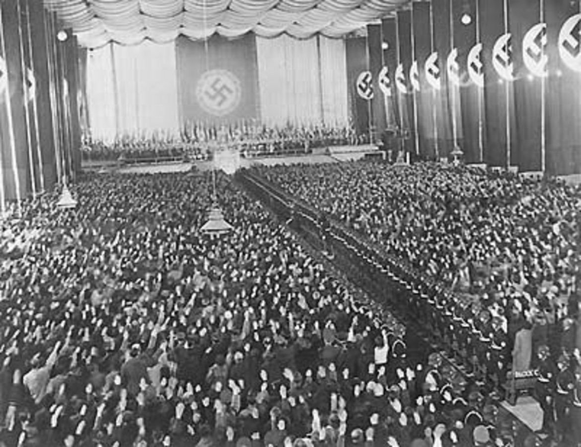 Das Schwarz-Weiß-Foto zeigt eine Menschenmenge in der Wiener Nordwesbahnhalle wo Adolf Hitler am 9. April 1938 zum Abschluss des Wahlkampf für die Volksabstimmung zum „Anschluss“ Österreichs an das Deutsche Reich eine Rede hält. Hinter der Bühne ist eine riesige Fahne mit Hakenkreuz zu sehen.