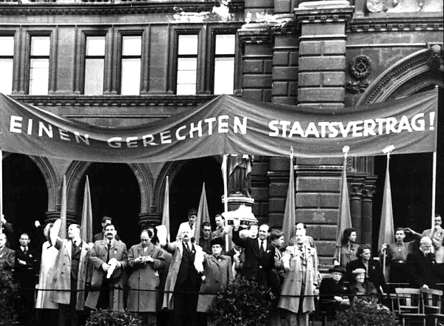 Auf einer Tribühne vor dem Rathaus stehen Frauen und Männer und winken. Davor Männer einer Blasmusikkapelle. Dahinter ein riesengroßes Transparent: "Wir fordern einen gerechten Staatsvertrag!"