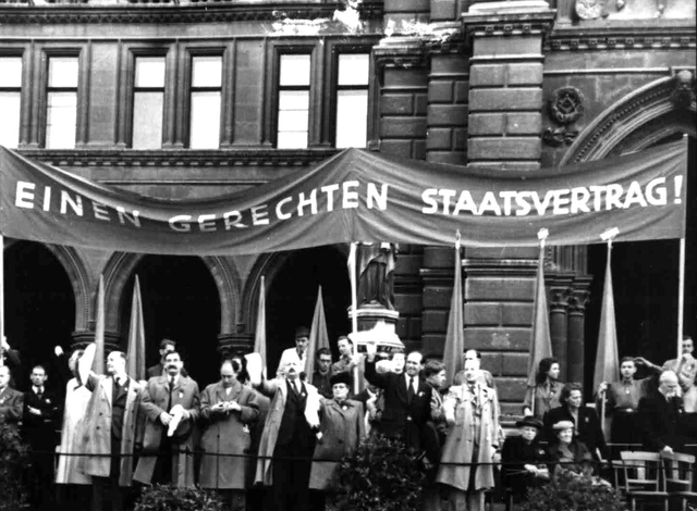 Auf einer Tribühne vor dem Rathaus stehen Frauen und Männer und winken. Davor Männer einer Blasmusikkapelle. Dahinter ein riesengroßes Transparent: "Wir fordern einen gerechten Staatsvertrag!"