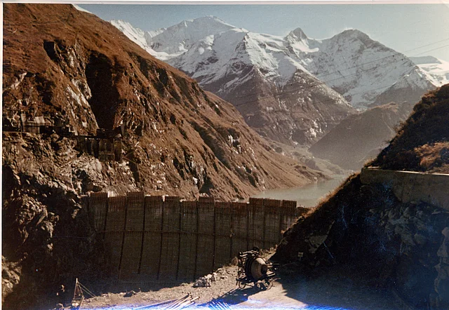 Farbfoto: Blick auf die noch nicht ganz fertiggestellte Staumauer vor Bergpanorama.