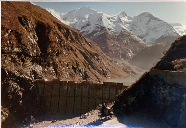 Farbfoto: Blick auf die noch nicht ganz fertiggestellte Staumauer vor Bergpanorama.