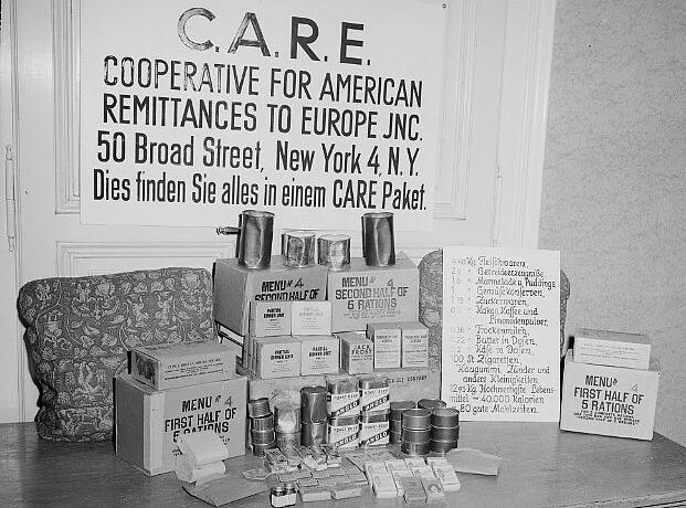 Der Inhalt eines CARE-Pakets wird am 9. Juli 1946 präsentiert. Fleischdosen, Marmelade, Pudding, Gemüsekonserven, Zucker, Kakao, Kaffee, Limonadenpulver, Trockenmilch, Butter, Käse, Zigaretten, Kaugummi ... Auf einem Tisch sind die kleinen Kisten und Dosen die sich in einem solchen Care-Paket befinden aufgestapelt. Dahinter ein Schild auf dem u.a. steht: C.A.R.E. Cooperative for American Remittances to Europe JNC.