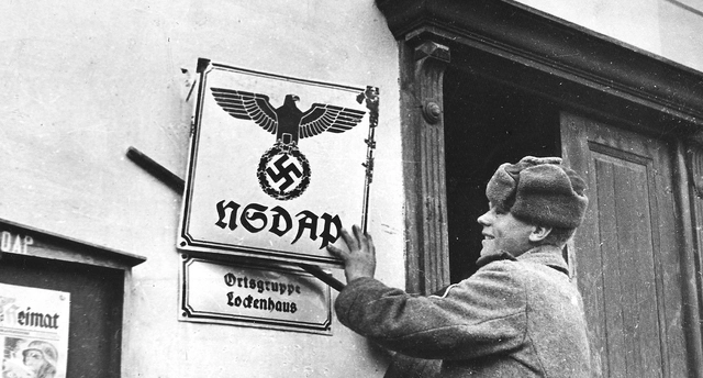 Soldat mit Fellmütze löst das NSDAP-Schild von der Hausmauer.