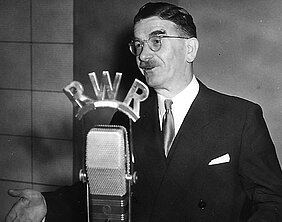 Leopold Figl vor dem Mirkofon über dem die Buchstaben RWR für Rot-Weiß-Rot angebracht sind.