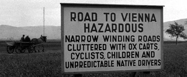 Großes Schild mit englischer Information: "ROAD TO VEINNA HAZARDOUS: NARROW WINDING ROADS CLUTTERED WITH OX CARTS; CYCISTS; CHILDREN AND UNPREDICTABLE NATIVE DRIVERS". Ein Pferdewagen im Hintergrund ist auf der Straße zu sehen.