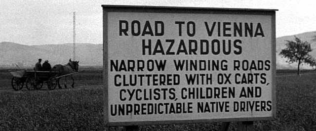 Großes Schild mit englischer Information: "ROAD TO VEINNA HAZARDOUS: NARROW WINDING ROADS CLUTTERED WITH OX CARTS; CYCISTS; CHILDREN AND UNPREDICTABLE NATIVE DRIVERS". Ein Pferdewagen im Hintergrund ist auf der Straße zu sehen.