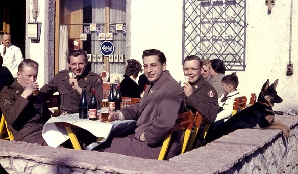 Farbfoto: 4 Soldaten an einem Tisch auf dem Bierflaschen und Biergläse stehen. Im Hintergrund andere Gäste sitzend und ein Kellner im weißen Sakko.