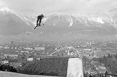 Blick vom Berg Isel auf die Stadt während des Skisprungbewerbs. Ein kleiner Skispringer ist vor dem Bergpanorama und dem Auslauf zu sehen.