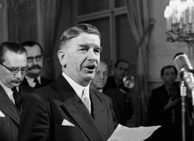 Leopold Figl mit Manuskriptblatt vor einen Mikrfon im Saal des Bunsekanzleramtes. Dahinter sind Männer in Anzügen sichtbar.