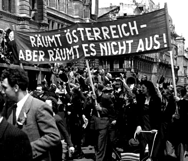 Links eine Holztribüne mit winkenden Politker_innen. Vorbeimarschierende Leute. Die Wiener Rathausfassade ist erkennbar und im Hintergrund sieht man die Votivkirche. Transparent: "Räumt Österreich - aber räumt es nicht aus!"