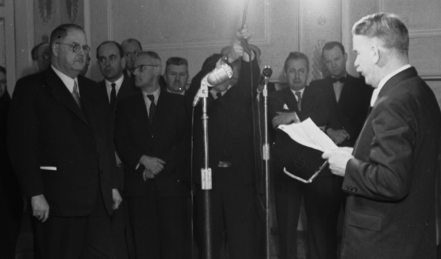 Außenminister Leopold Figl vor dem Mikrofon spricht zu dem gegenüber stehenden Julius Raab. Hinter diesem Männer in Anzügen die zuhören.
