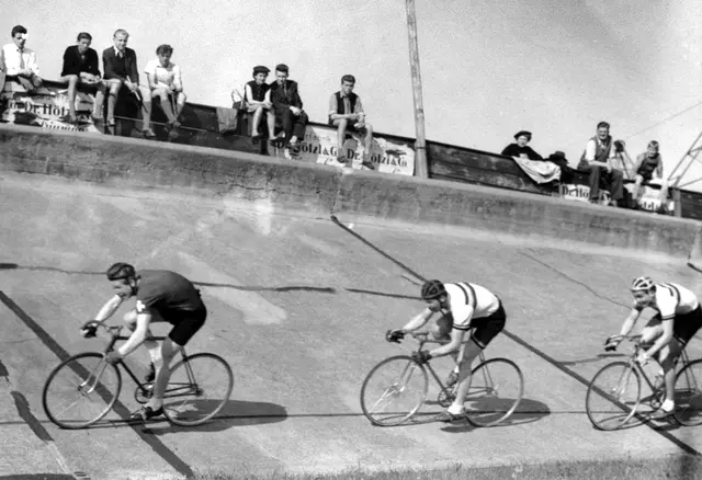 Drei Radfahrer im Freilichtradstadion. An der Oberkante der Steilkurfe sind einige Zuschauer zu sehen.