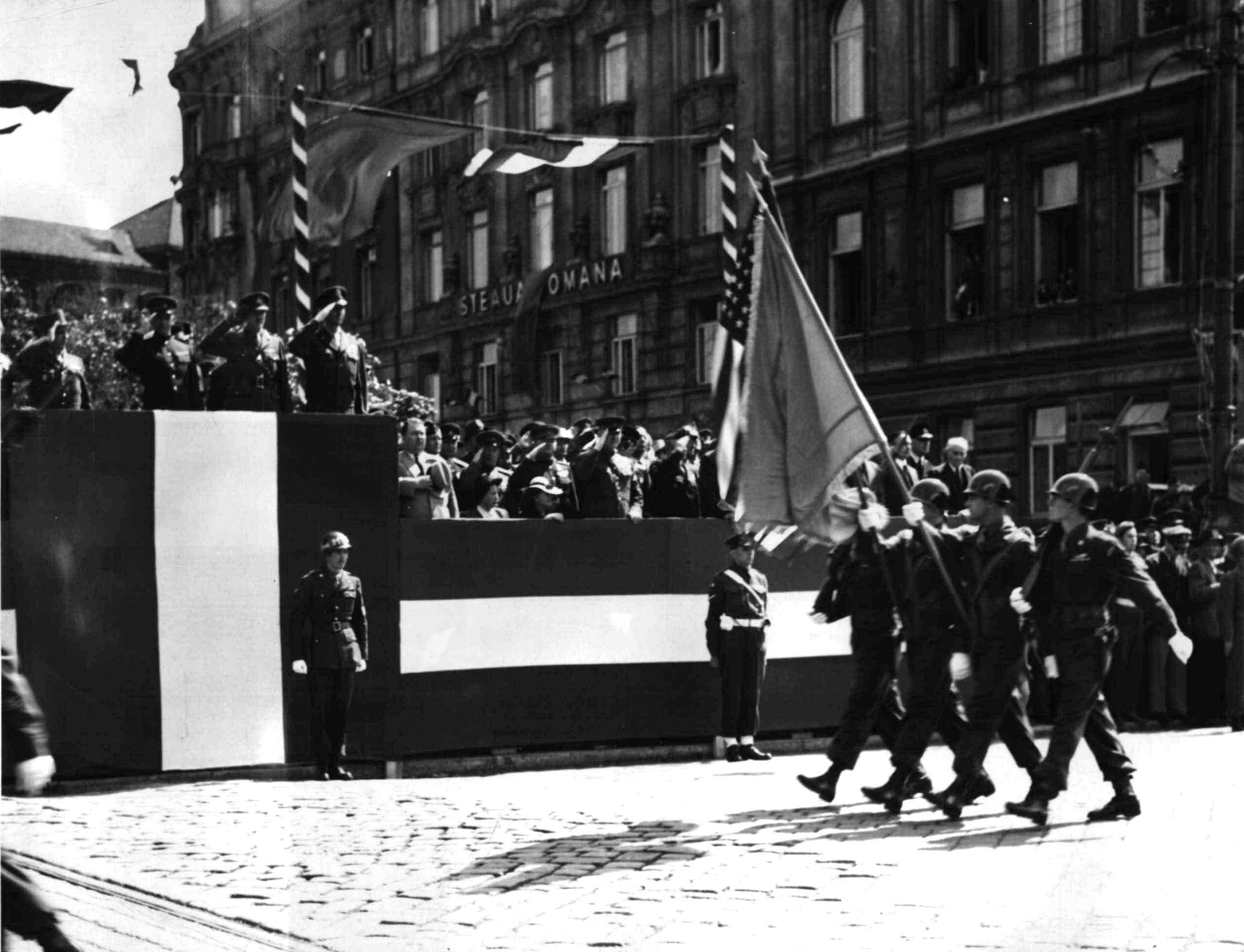 Salutierende Kommandeure auf rot-weiss-rot verkleideten Tribünen nehmen ein Militärparade ab. Hier sind vier US-amerikanische Soldaten mit Stahlhelmen - zwei mit geschulterten Gewehren und zwei mit Flaggen zu erkennen.