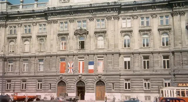 Blick auf das Gebäude mit einigen Autos im Vordergrund. Die Flaggen der Alliierten und Österreichs sind über dem Eingang nebeneinander aufgespannt.
