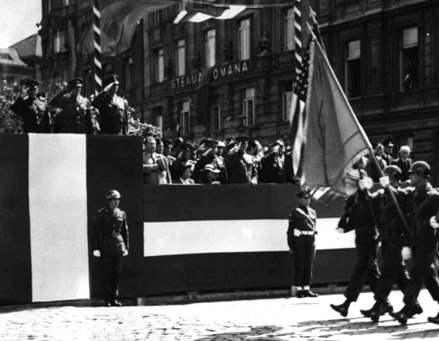 Salutierende Kommandeure auf rot-weiss-rot verkleideten Tribünen nehmen ein Militärparade ab. Hier sind vier US-amerikanische Soldaten mit Stahlhelmen - zwei mit geschulterten Gewehren und zwei mit Flaggen zu erkennen.