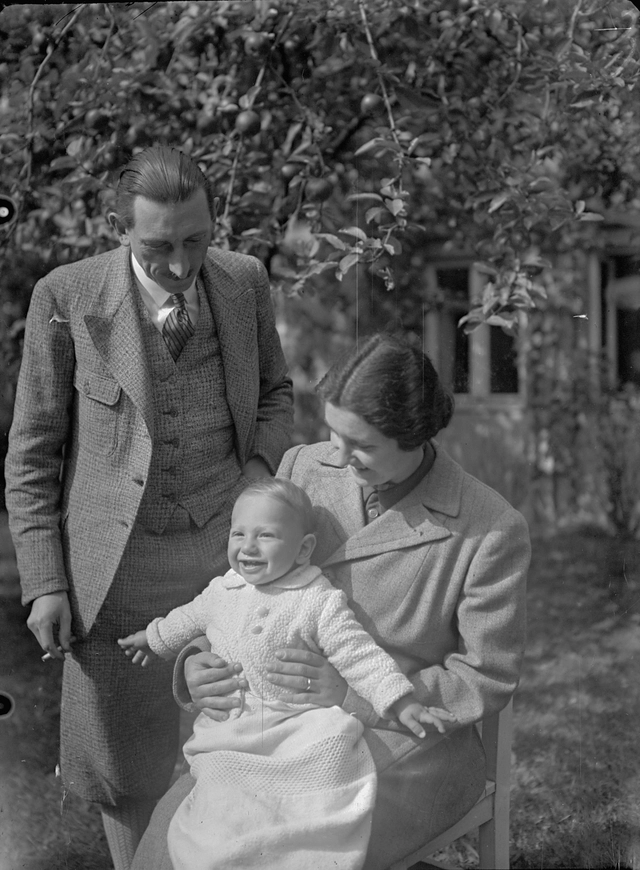 Ein elegant gekleideter Herr steht neben einer Dame im Mantel, die sitzend ein Baby am Schoß hält. Beide Erwachsenen schauen auf das Baby. Im Hintergrund ist ein Apfelbaum, ein Garten und ein Haus sichtbar.(schwarzweiß)