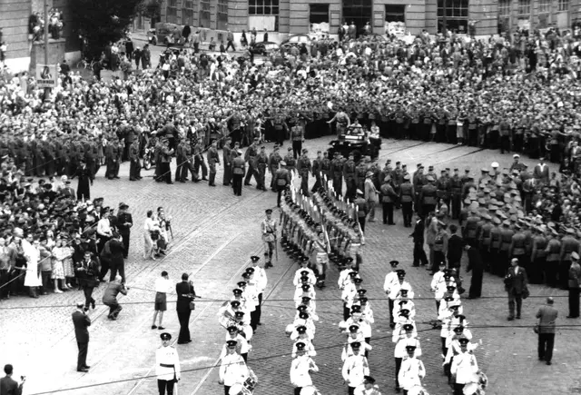 Viele Soldaten in Formation, sowie eine Militärkapelle aus erhöhten Standpunkt fotografiert. Viele Schaulustige sind an den Rändern zu sehen.