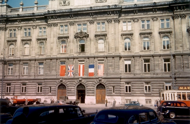 Blick auf das Gebäude mit einigen Autos im Vordergrund. Die Flaggen der Alliierten und Österreichs sind über dem Eingang nebeneinander aufgespannt.