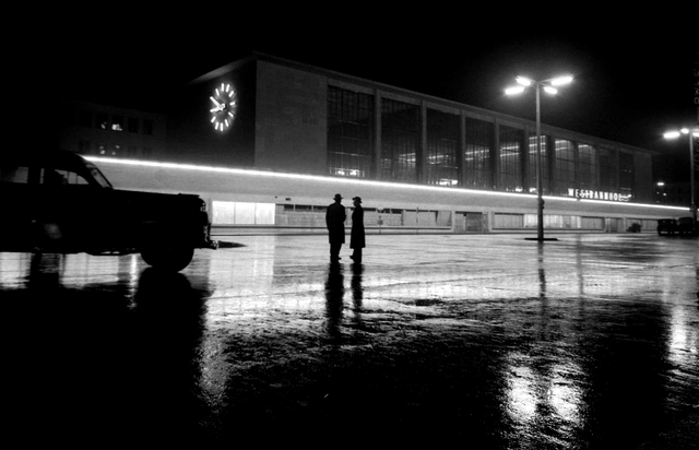 Hellspiegelnder nasser Asphat. Siluette eines PKWs und von zwei Männern mit Mantel und Hut vor dem modernen Gebäude des Westbahnhofs. Straßenlaternen und die Uhr des Bahnhofs (10 vor 9 Uhr) leuchten.