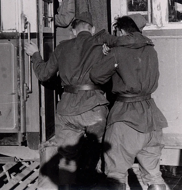 Rückenansicht der beiden Soldaten. Der eine hilft dem anderen - schwer beeinträchtigten - in die Straßenbahn hinauf.