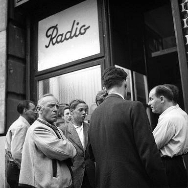 Männer hören vor einem Radiogeschäft mit ernster Mine der Radioreportage zu.