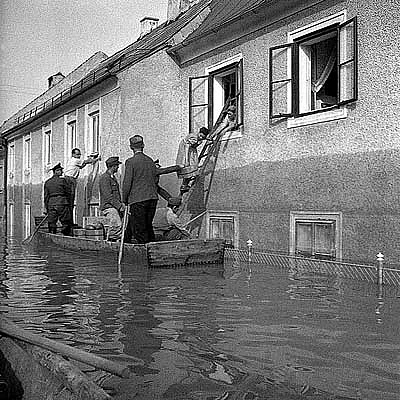 Feuerwehrleute sind mit Zillen unterwegs und betreuen die Flutopfer, an die sie amerikanische CARE-Pakete verteilen. Linz, 14. Juli 1954. Eine Frau übernimmt ein Paket und klettert auf einer Leiter durchs Fenster der Zille entgegen.