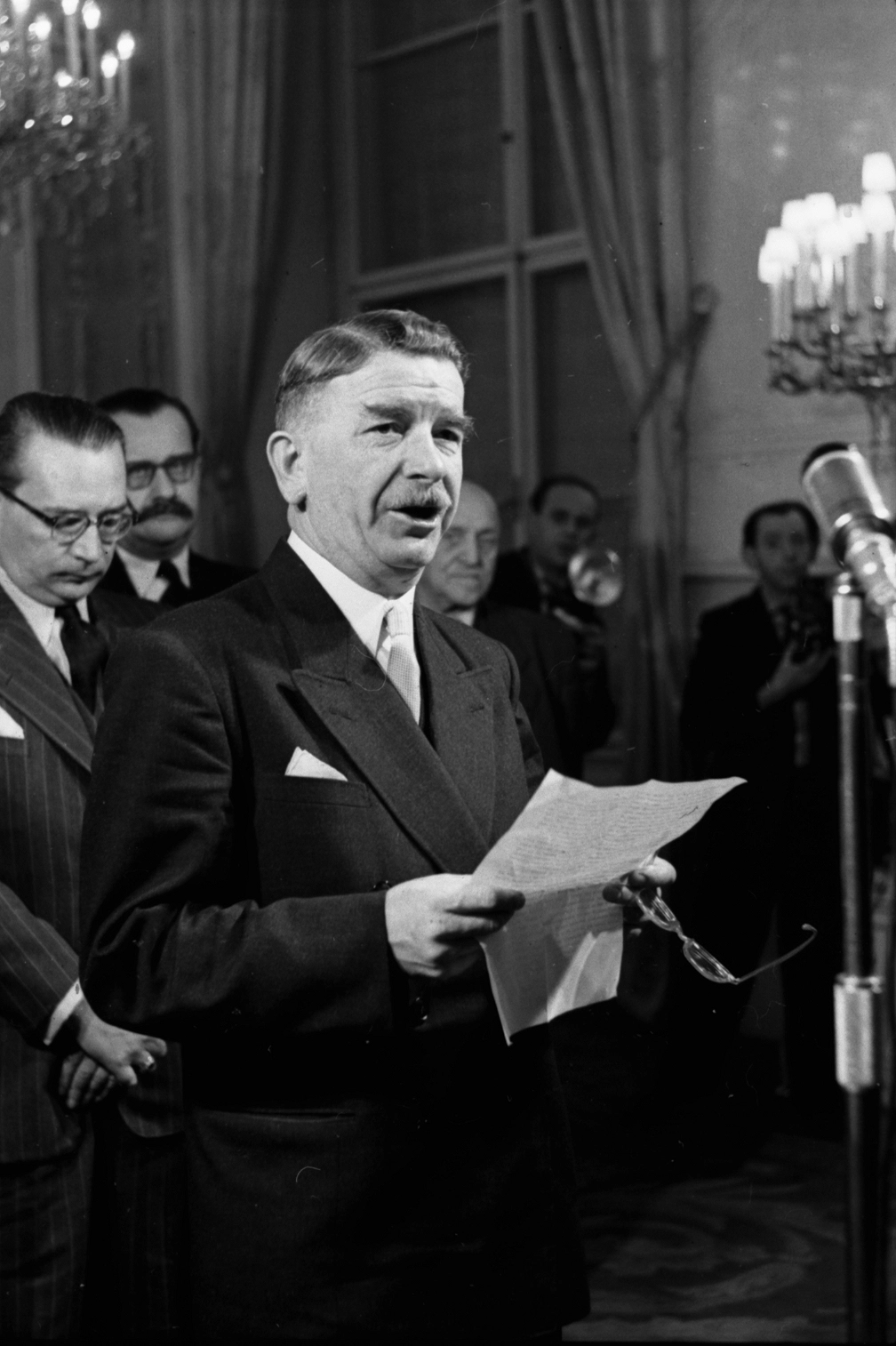 Leopold Figl mit Manuskriptblatt vor einen Mikrfon im Saal des Bunsekanzleramtes. Dahinter sind Männer in Anzügen sichtbar.