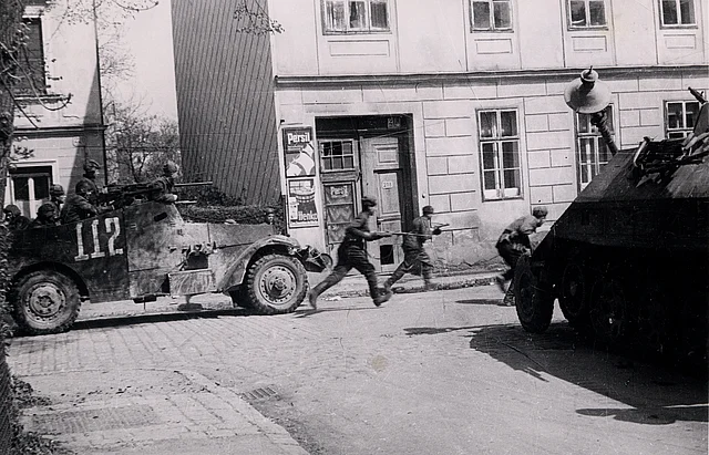 Soldaten zu Fuß und im Militärfahrzeug in einer Straße in Wien. Ein Haus mit Plakaten neben dem Hauseingang ist zu erkennen.