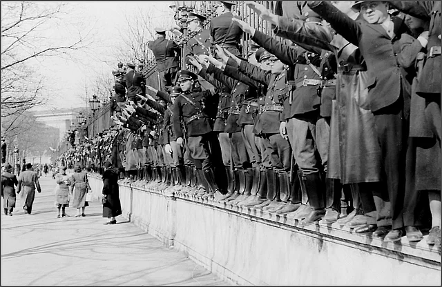 Auf der Umfriedung des Heldenplatzes drängen sich viele Menschen, darunter auch zahlreiche Polizisten, die den rechten Arm zum Hitlergruß erhoben haben.