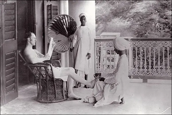 Ein Brite in Indien, dem von einem Diener die Füsse gewaschen werden, während ein zweiter Diener für Kühlung mit einem Fächer sorgt.