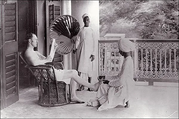 Ein Brite in Indien, dem von einem Diener die Füsse gewaschen werden, während ein zweiter Diener für Kühlung mit einem Fächer sorgt.