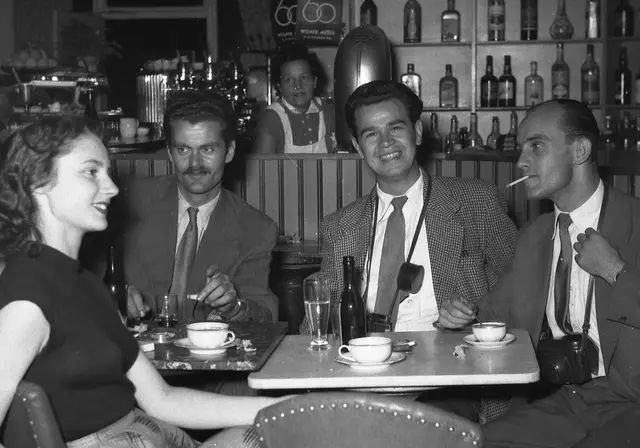 Drei Männer - teilweise mit umgehängten Kameras - und eine Frau an einem Kaffeehaustisch. Im Hintergrund ist die Bar zu sehen.