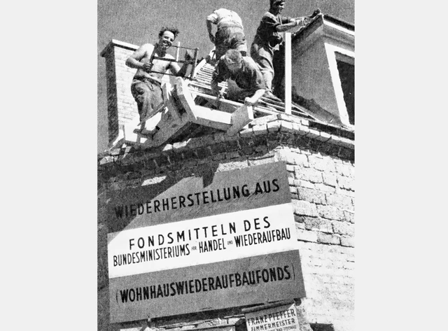 Vier Arbeiter werken auf einem Dachstuhl. Darunter das Plakat: Wiederherstellung aus Fondsmitteln des Bundesministeriums für Handel und Wiederaufbau (Wohnhauswiederaufbaufonds)"