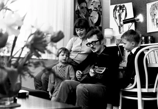 Vierköpfige Familie. Der Vater spielt Gitarre, Mutter und Sohn und Tochter sitzen dabei im Wohnzimmer mit Bildern an den Wänden.
