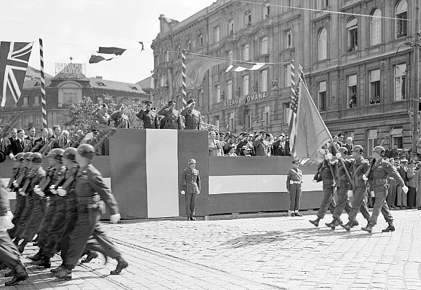 Salutierende Kommandeure auf rot-weiss-rot verkleideten Tribünen nehmen ein Militärparade ab. Hier sind vier US-amerikanische Soldaten mit Stahlhelmen - zwei mit geschulterten Gewehren und zwei mit Flaggen zu erkennen. Ganz links marschieren Soldaten aus dem Bild.