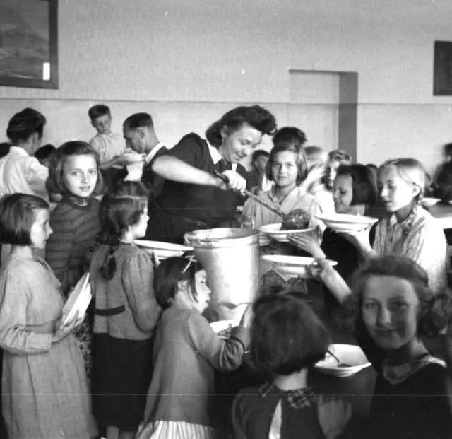 In einer Schulklasse gibt eine Frau mit Schöpfer Suppe aus. Kinder umringen sie mit Tellern und warten.