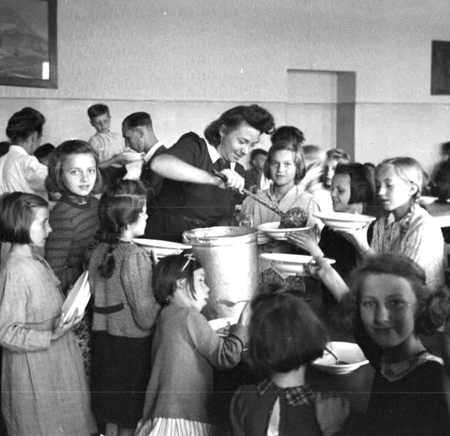 In einer Schulklasse gibt eine Frau mit Schöpfer Suppe aus. Kinder umringen sie mit Tellern und warten.