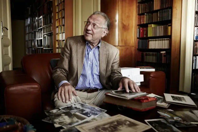 Gerhard Jagschitz sitzt auf einem Ledersessel und schaut nach links. Vor ihm liegen historische Fotos auf einem Couchtisch. Im Hintergrund ist ein Bücherregal sichtbar.