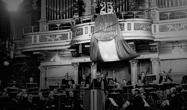 Festsaal mit Orchester auf der Bühne sowie einem Redner hinter einem Podium sichtbar. 