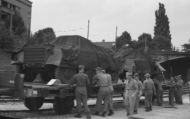 US-amerikanische Soldaten beim Aufladen von Spähpanzern. Soldaten umringen einen einen offenen Eisenbahnwaggon auf dem zwei Panzerwägen unter Planen aufgeladen wurden und scheinen die Ladung zu kontrollieren.