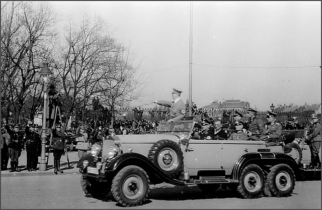 Auf der Schwarz-Weiß-Fotografie sieht man Adolf Hitler mit erhobenem rechtem Arm, wie er in seinem Auto an der Menschenmenge vorbeifährt.