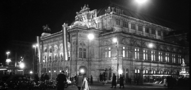 Nachtaufnahme der beleuchteten und beflaggten Staatsoper. Einge Paare strömen über dem Ring zum Gebäude.