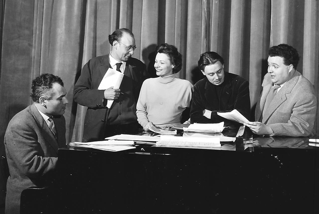 Von links: Jeff Palme, Carl Merz, Hilde Krahl, Otto Zokan (=Otto Kobalek) und Helmut Qualtinger. Mann (ganz links) am Klavier (Flügel) sitzend. Vier Personen stehen dahinter und schauen sich an oder in Manuskripte, die zusätzlich auch auch vor ihnen am Flügel liegen.