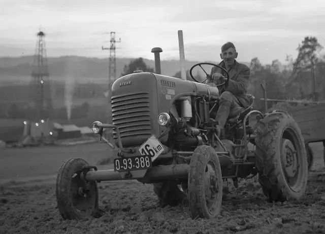 Ein Bauer auf einem Steyr-Traktor. Im Hintergrund ein Bohrturm und ein Hochspannungsmast.