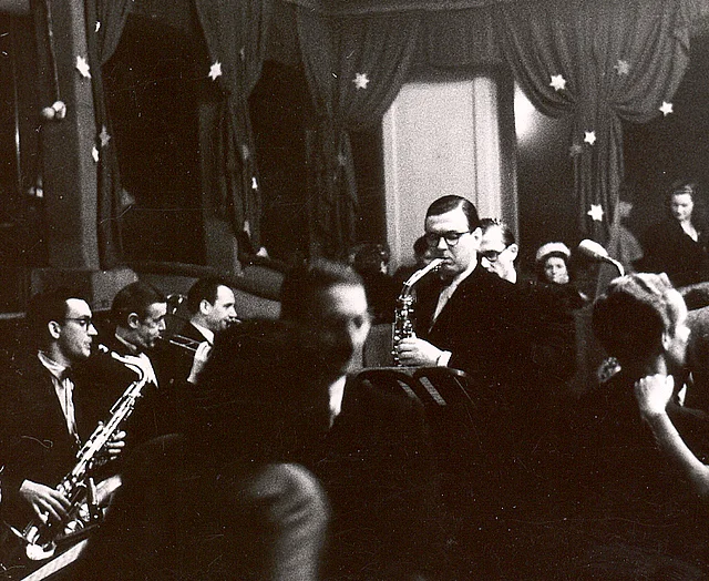 Links von ihm seine Musiker, auch mit Saxophonen, vor ihm und rechts sind Tanzpaare zu sehen.