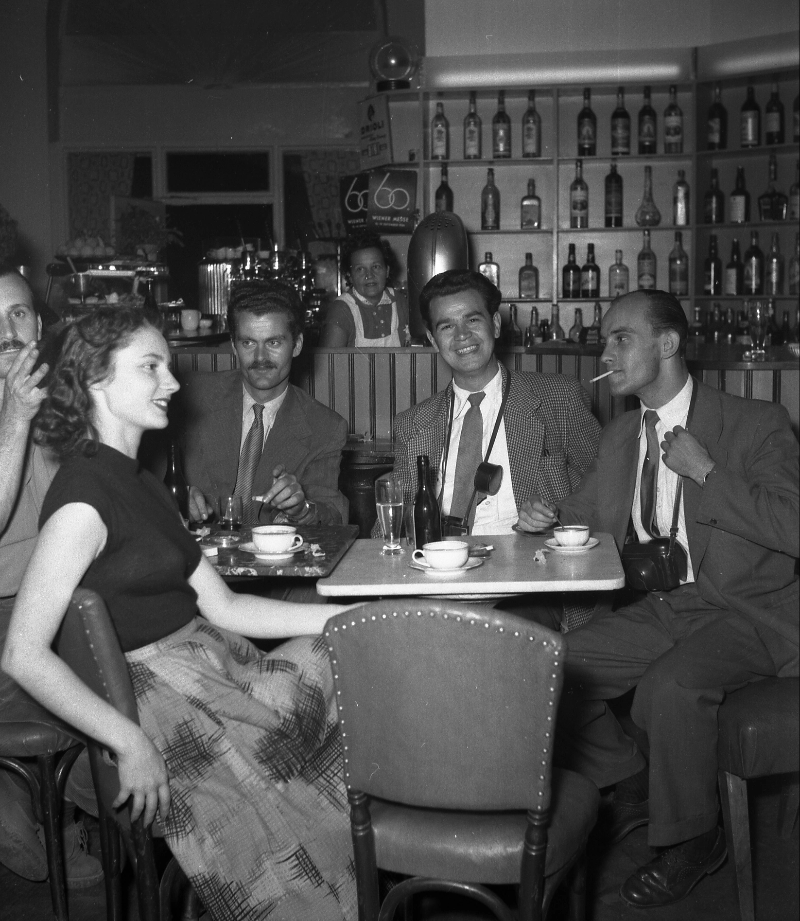 Drei Männer - teilweise mit umgehängten Kameras - und eine Frau an einem Kaffeehaustisch. Im Hintergrund ist die Bar zu sehen.