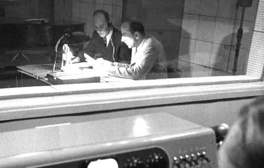 Zoom in das Radiostudio wo zwei Männer an einem Tisch Manuskripte lesen.