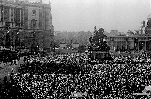 Blick aus einem Fenster der Neuen Hofburg auf die Menschenmenge am Heldenplatz. Auch das Reiterdenkal von Prinz Eugen ist zu sehen. Im Hintergrund sieht man das mit Hakenkreuzfahnen geschmückte Burgtor.