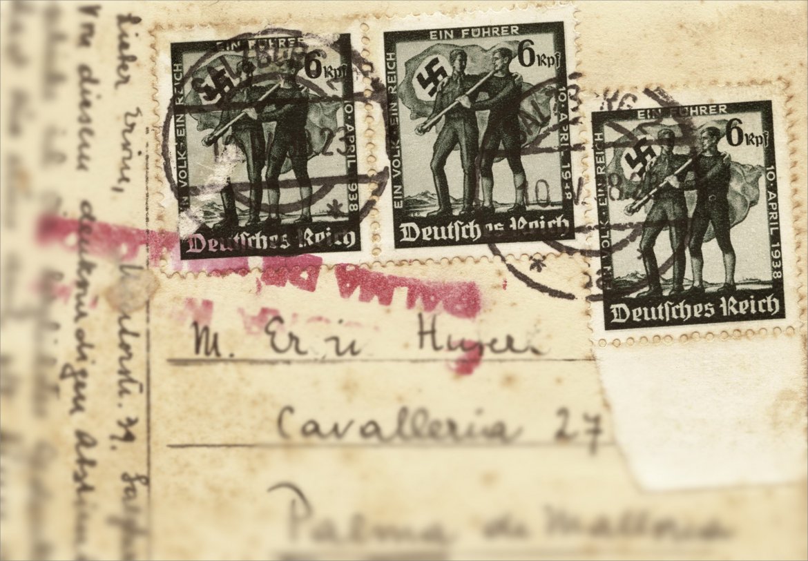 Zusehen ist eine Postkarte mit drei Briefmarken, die den „Anschluss“ propagieren. Am Rand der Marke steht "Ein Volk - Ein Reich - Ein Führer - 10. April 1938" in der Mitte sind zwei Männer zu sehen, die sich die Arme um die Schultern legen und eine große Hakenkreuzfahne tragen.
