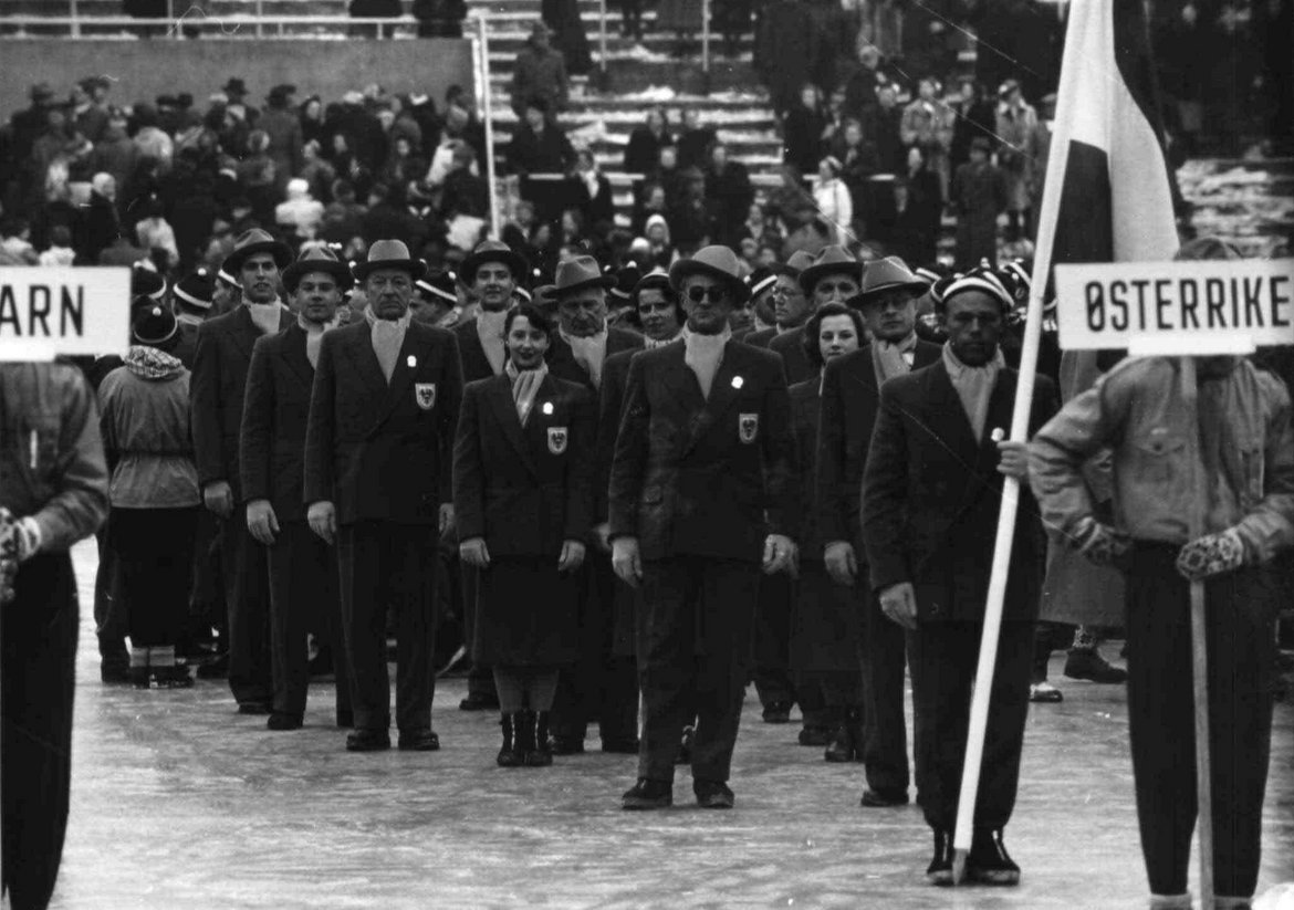 Gruppe von Sportlerinnen und Sportlern in Anzügen mit Hut und dem Fahnenträger, sowie einem Träger einer Tafel mit der Aufschrift"Osterrike"